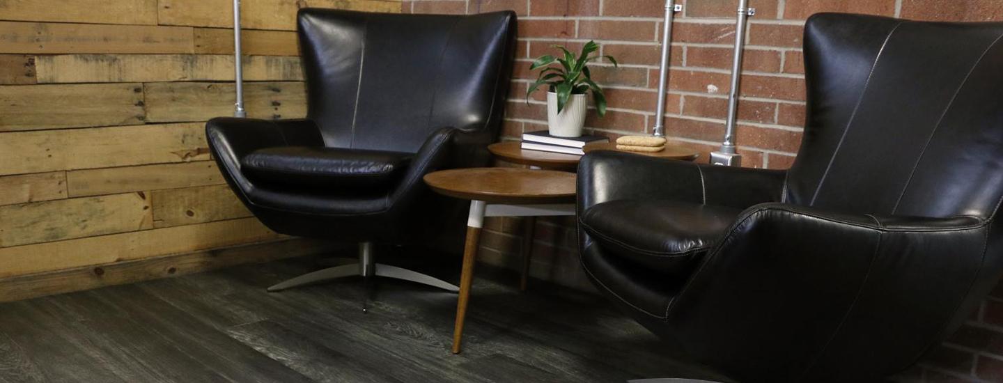 Stylish office space with two black leather chairs, a wooden table with books and a plant, against a rustic wood and brick wall.