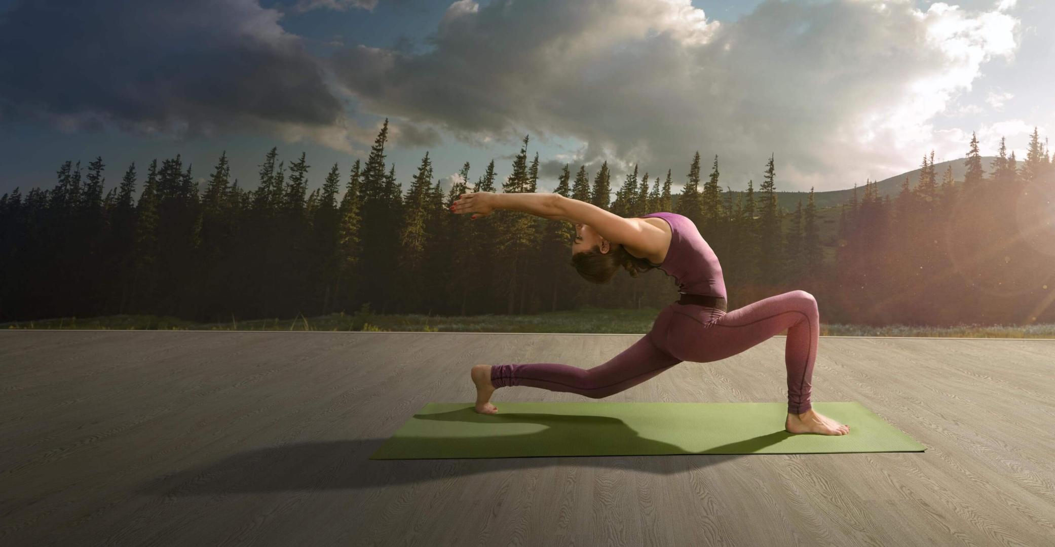 Woman practicing a yoga pose on a green mat outdoors, with a serene forest and mountain landscape in the background.