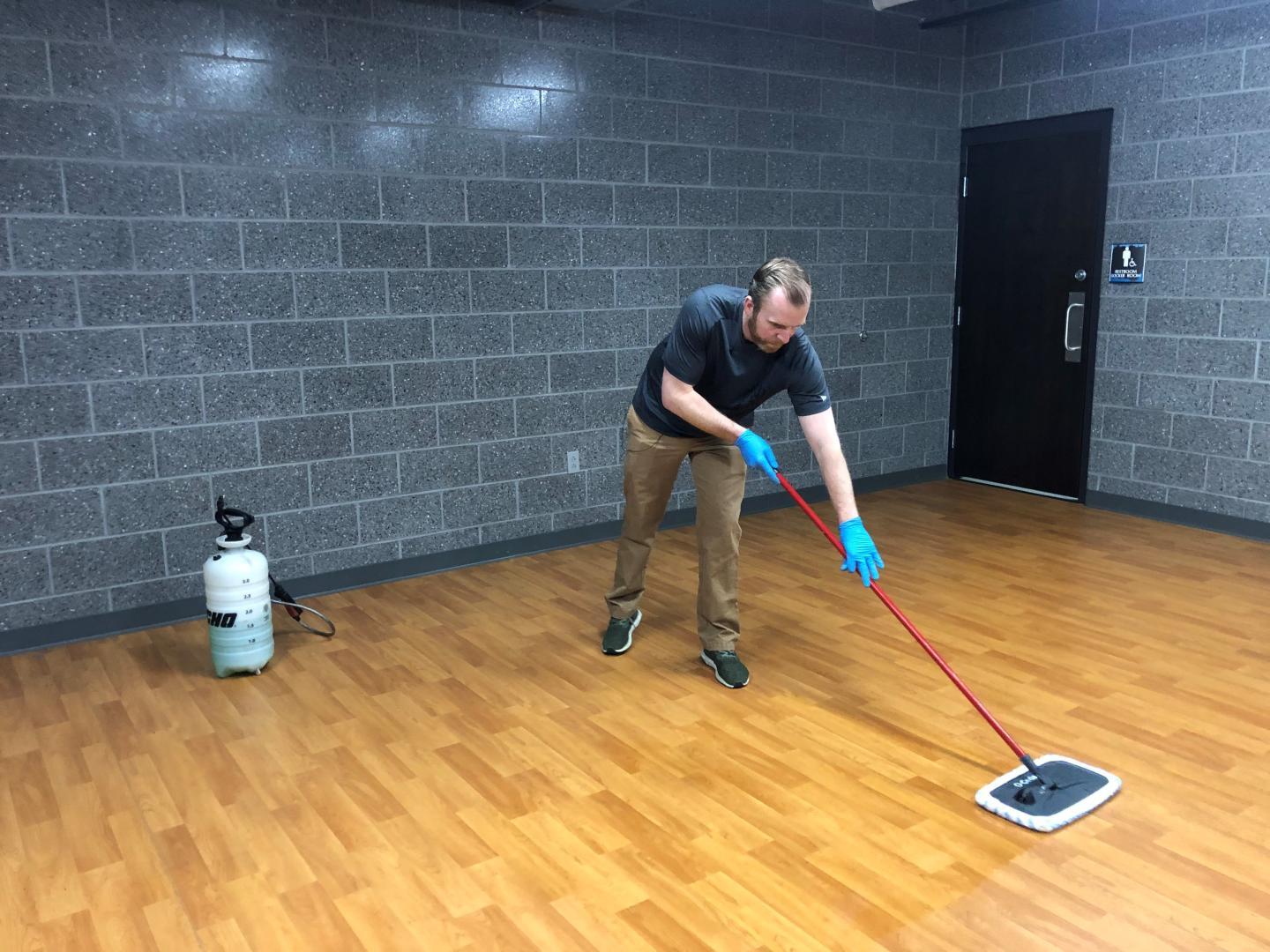 Man wearing janitorial gloves cleaning an indoor sports floor with cleaning product and sweeper mop.