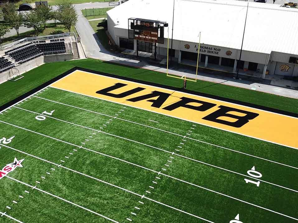 Aerial view of a football field with 'UAPB' in large black letters on the end zone, adjacent to stadium seating and a field house.