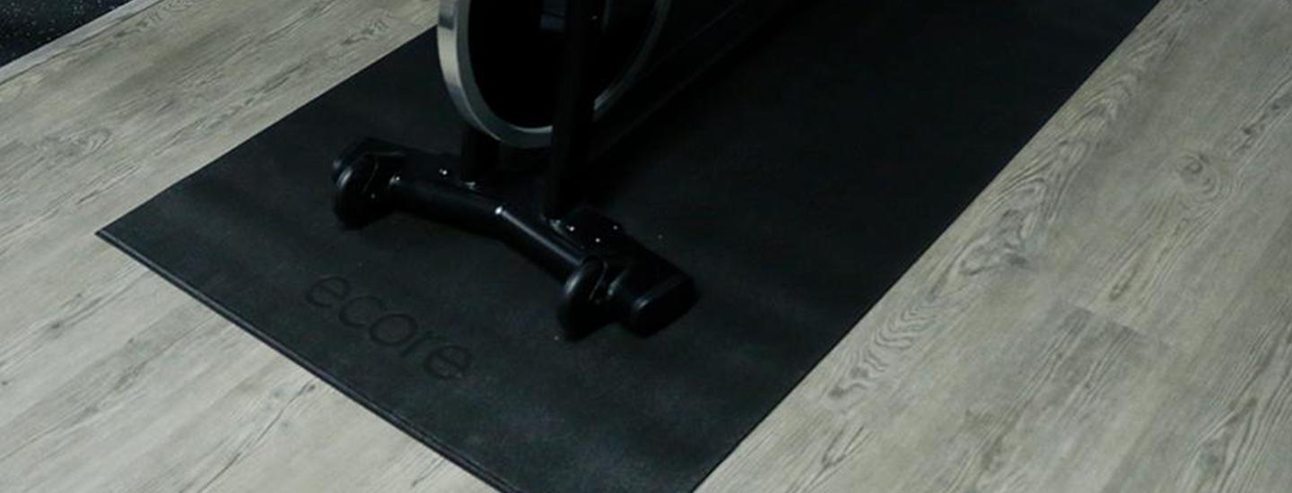 Close-up of a black gym floor mat branded 'ecore' with exercise equipment and gray flooring. 